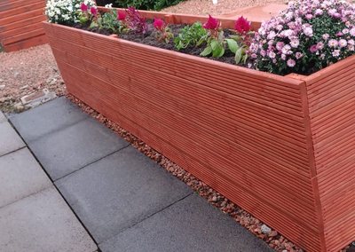 Stained decking-board planter with flowers next to a hedge